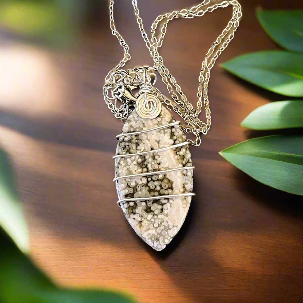 Green & White Ocean Geode Jasper Wire Wrapped Marquise Natural Stone Amulet Silver Necklace on wood table with leaves around the edges as decoration.